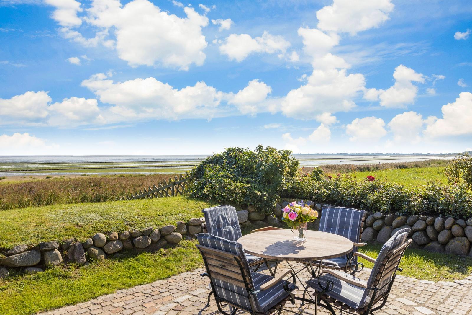 Terrasse mit Tisch und Stühlen, Blick auf Wiesen und Meer unter blauem Himmel.