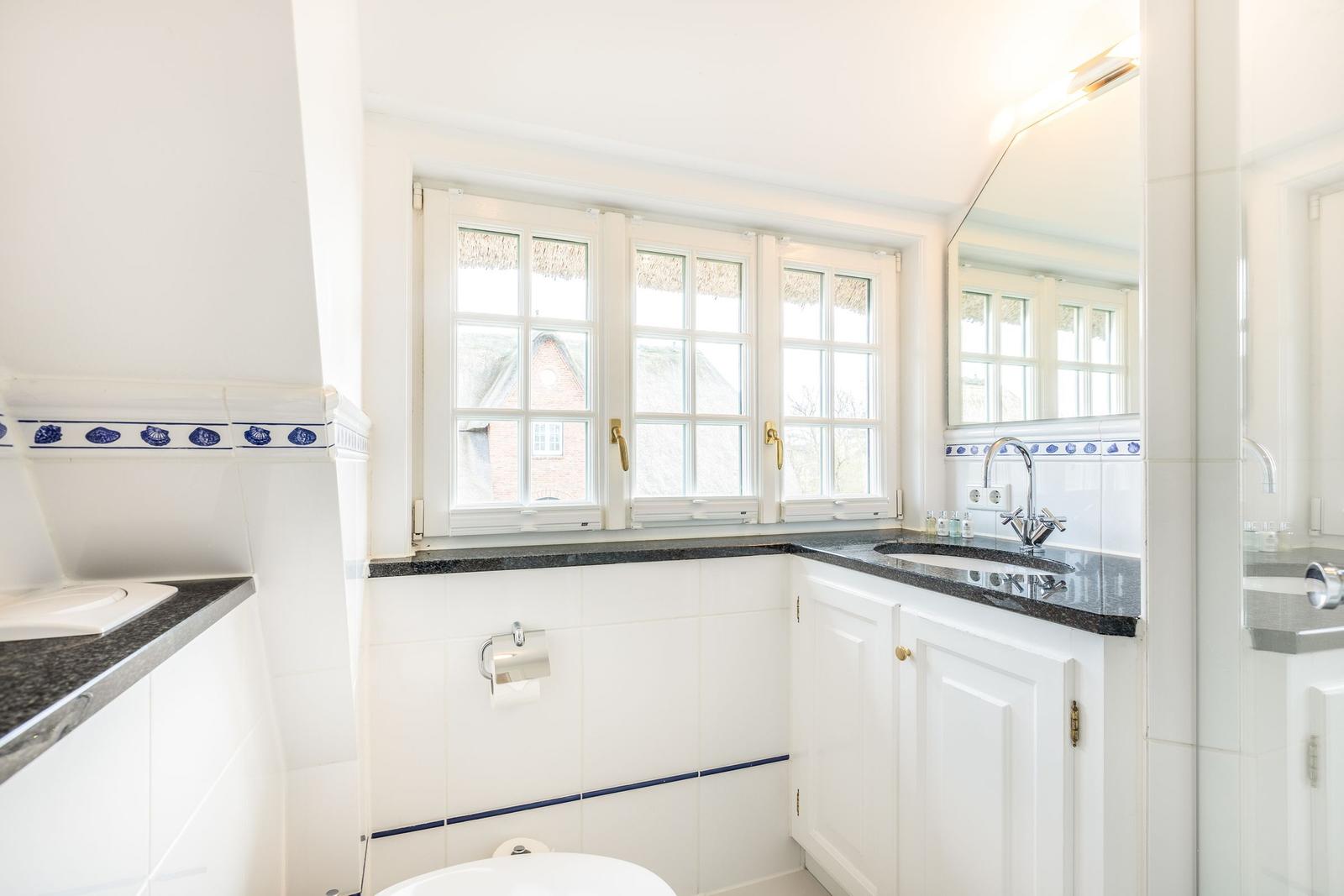 Badezimmer mit Fenster, Waschbecken und Toilette. Weiße Fliesen und dunkler Arbeitsplatz.