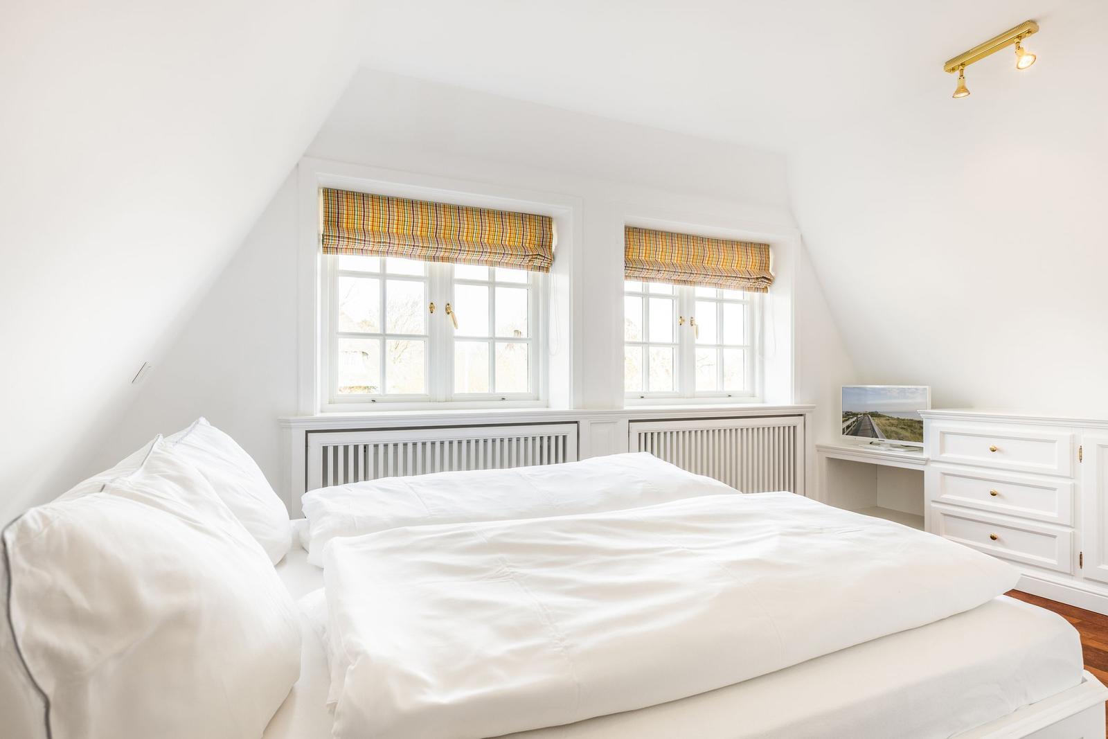 Schlafzimmer mit Dachschräge, zwei Fenstern und weißem Bett.