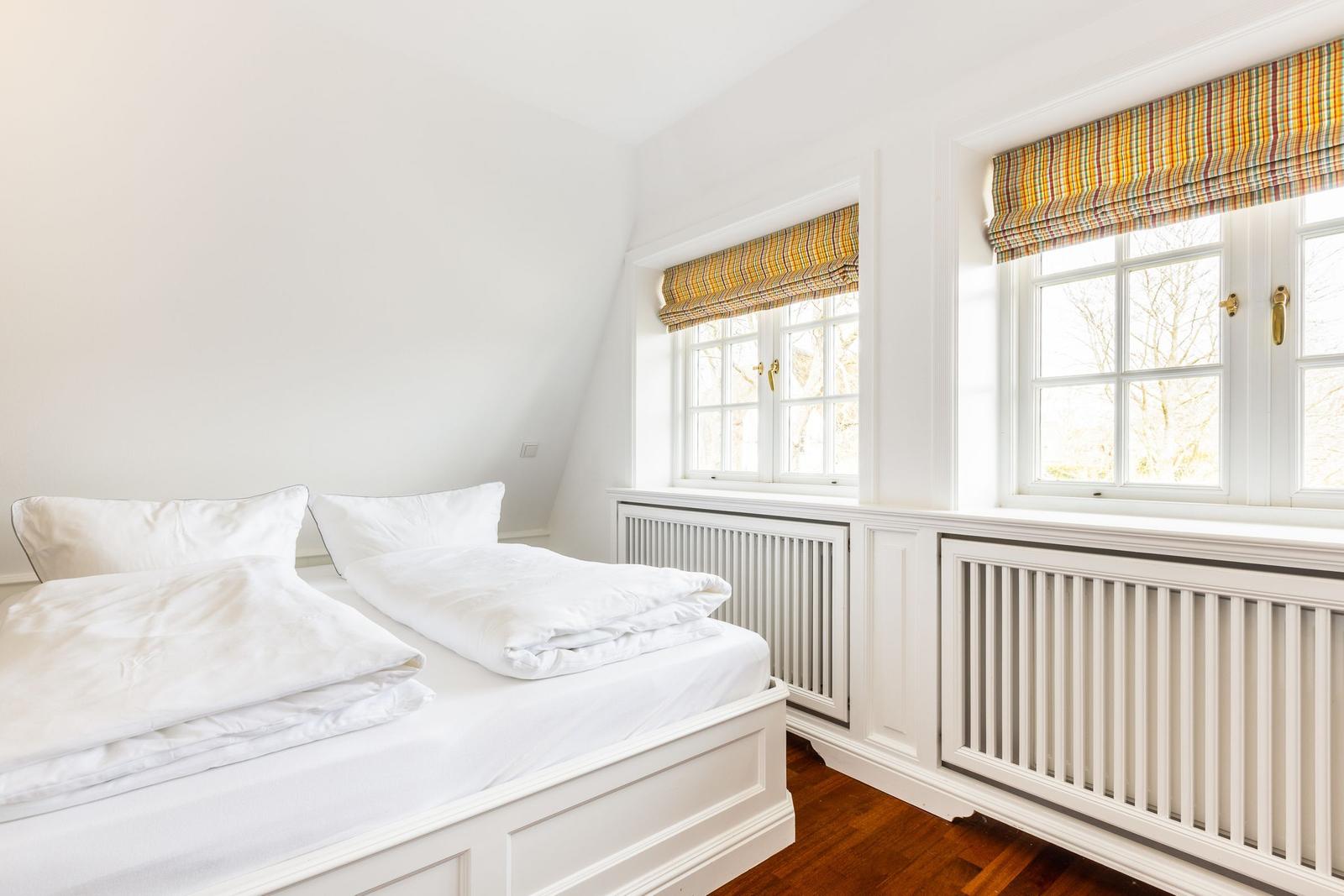 Schlafzimmer mit Doppelbett, weißen Wänden und zwei Fenstern mit bunten Rollläden.