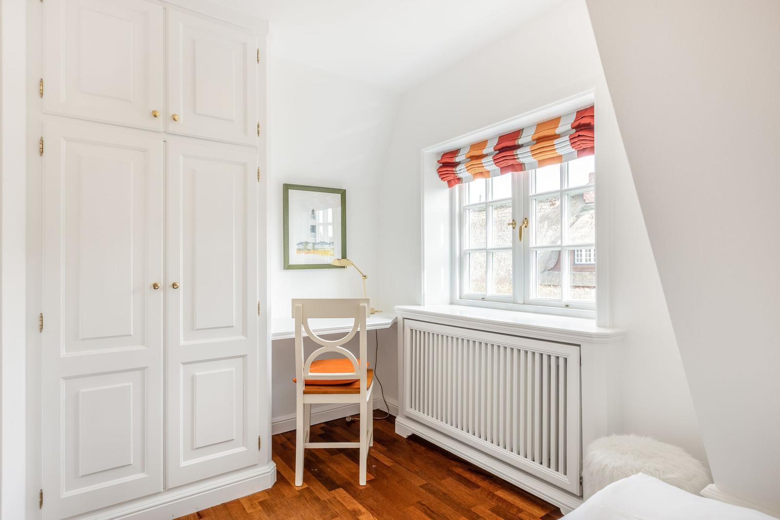 Schlafzimmer mit weißem Schrank, Schreibtisch und Fenster mit buntem Rollladen.