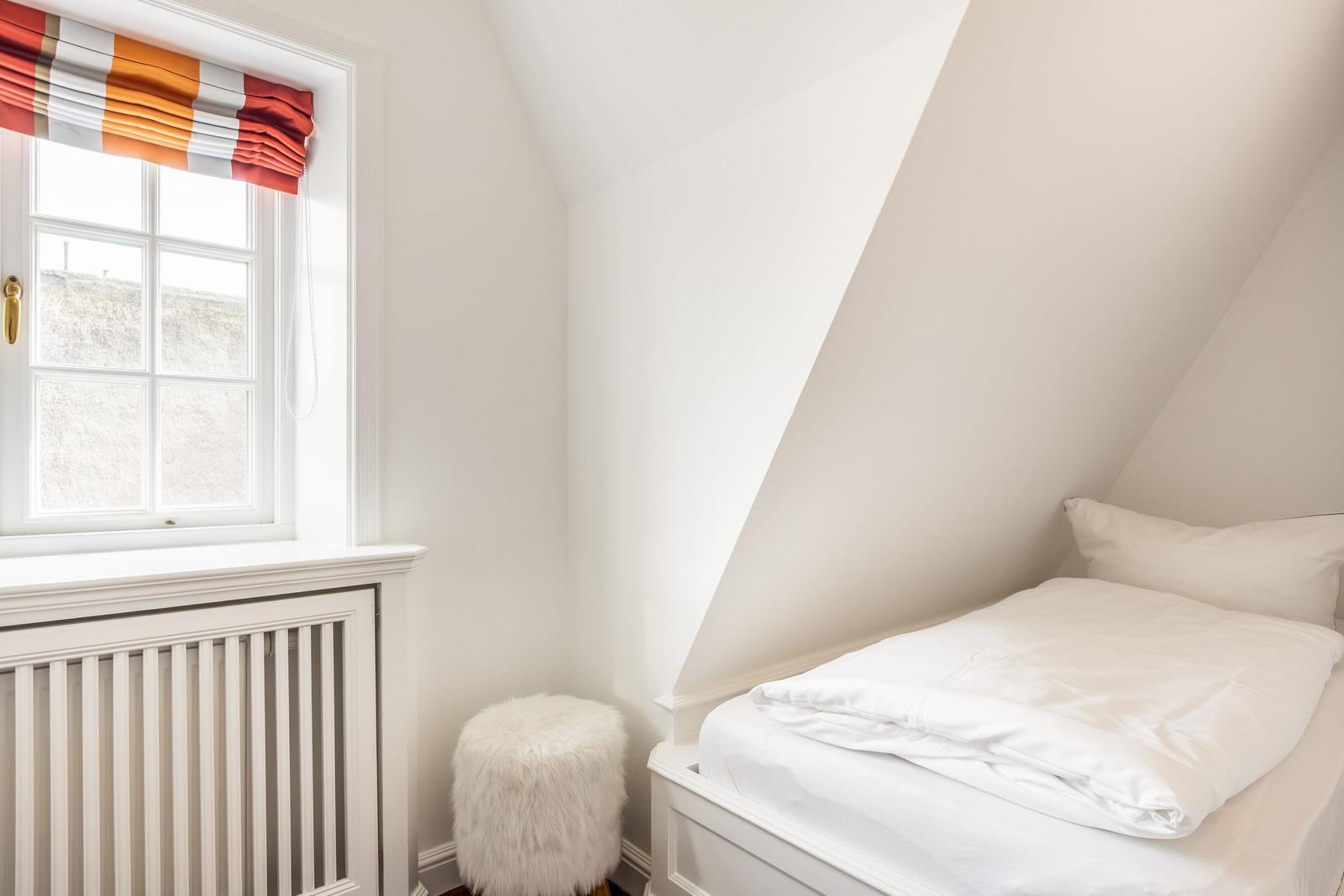 Ein Schlafzimmer mit weißem Bett, Fenster mit Rollladen und Fellhocker.