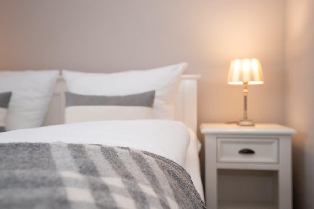 Ein Bett mit weißen Kissen und einer grau-weißen Decke neben einem Nachttisch mit Lampe.