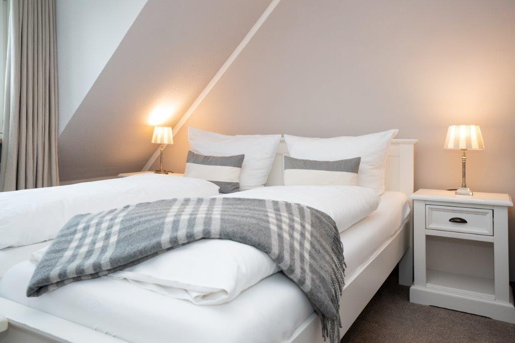 Schlafzimmer mit Dachschräge, Bett mit weißen Laken und grauer Decke, zwei Nachttische mit Lampen.
