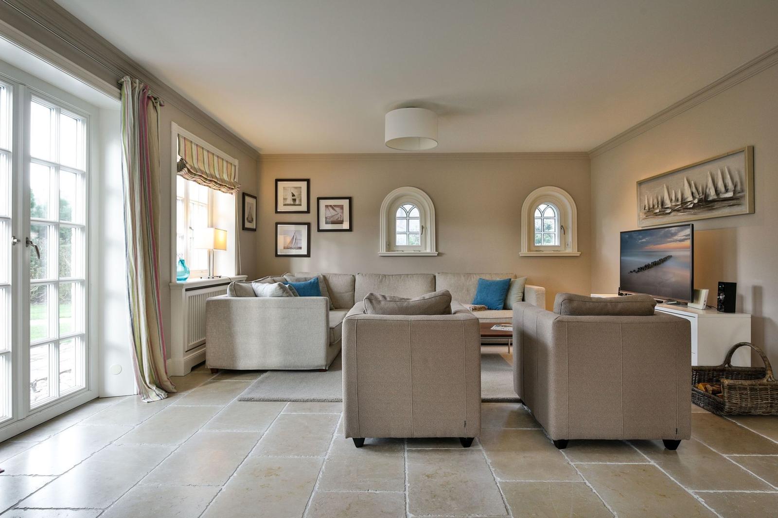 Wohnzimmer mit Sofa, Sesseln, TV und Steinboden. Fenster und Wandbilder sichtbar.