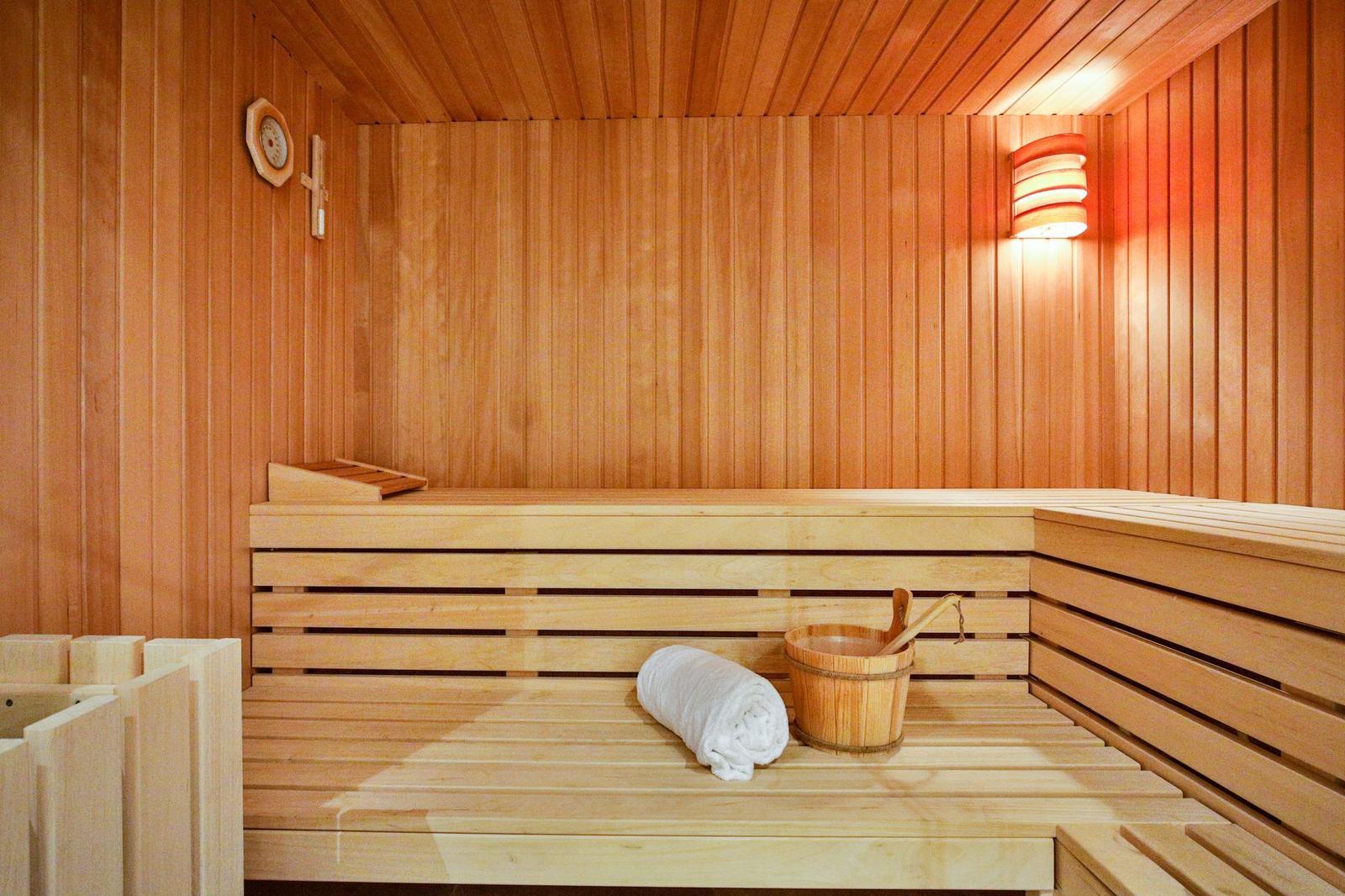 Sauna mit Holzbank, weißem Handtuch und Eimer. Wanduhr und Wandlampe sichtbar.