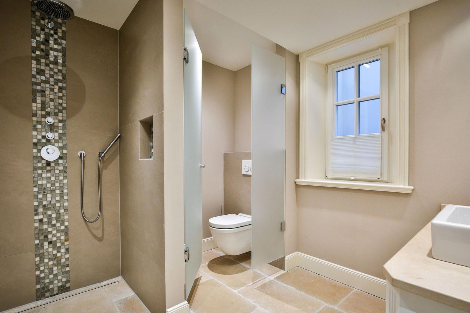Dusche mit Mosaikfliesen, Toilette hinter Glaswand und Waschtisch mit Fenster.