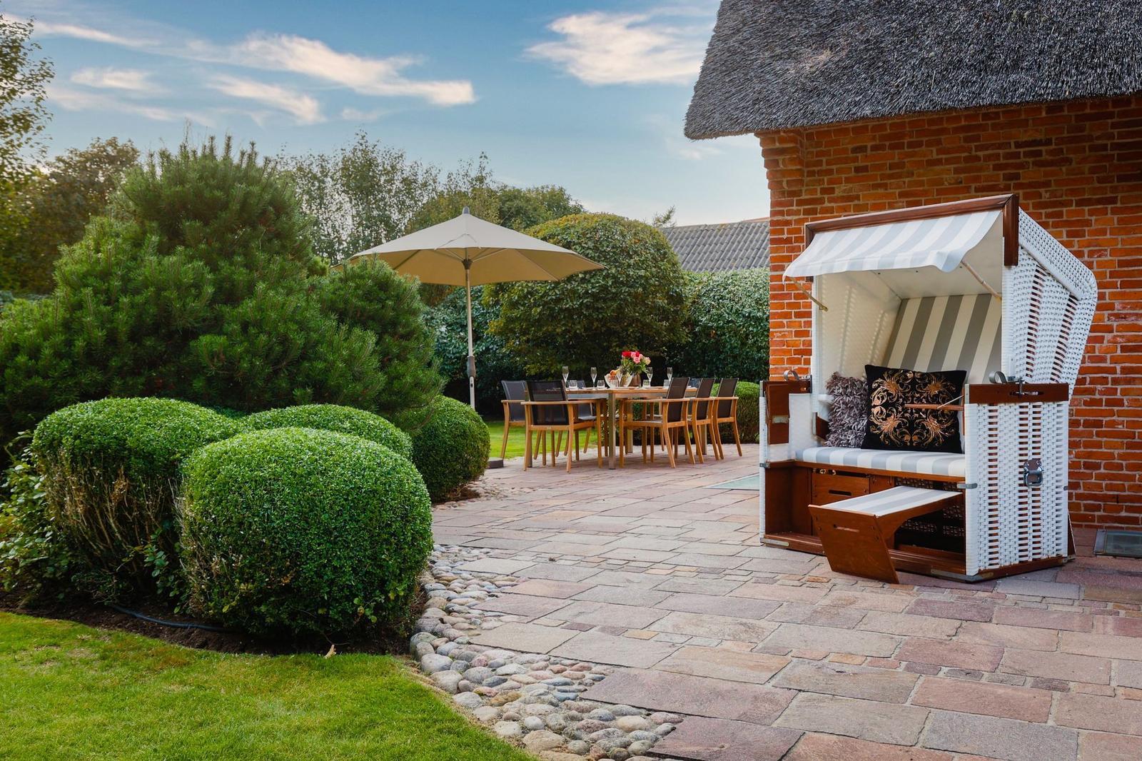 Terrasse mit Strandkorb, Tisch und Stühlen, umgeben von grünen Büschen und einem Steinweg.