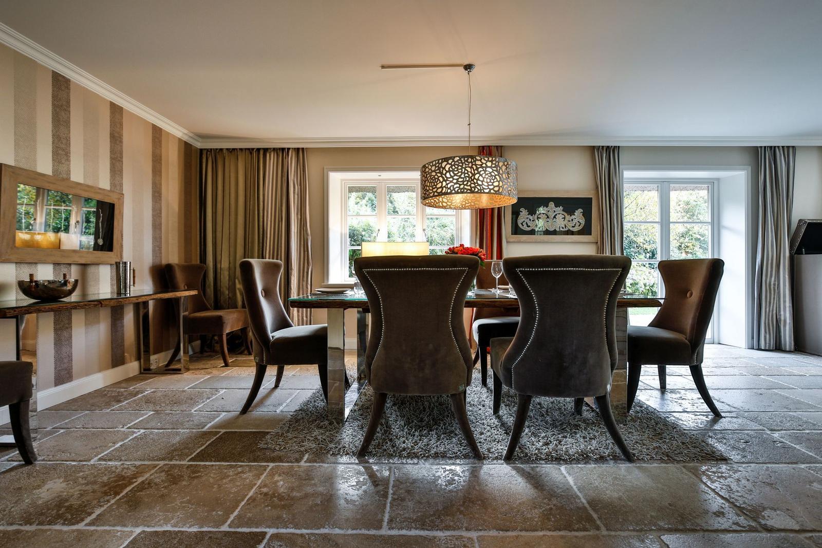 Esszimmer mit Holztisch, Stühlen und Steinboden. Fenster mit Vorhängen und Deckenleuchte.