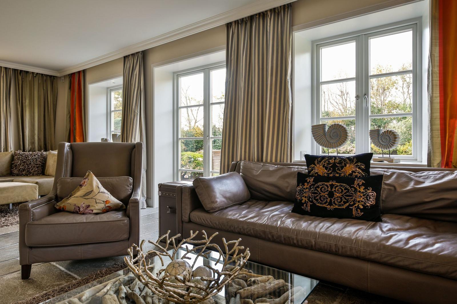 Wohnzimmer mit Ledersofa, Sessel und Glas-Tisch. Fenster mit Gardinen und Dekoration.