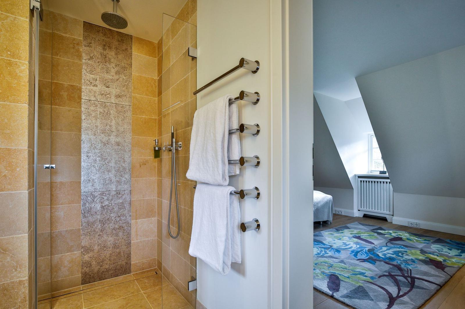 Dusche mit Glaswand und Handbrause. Badetuchhalter mit weißen Handtüchern. Blick in Schlafzimmer mit Teppich.