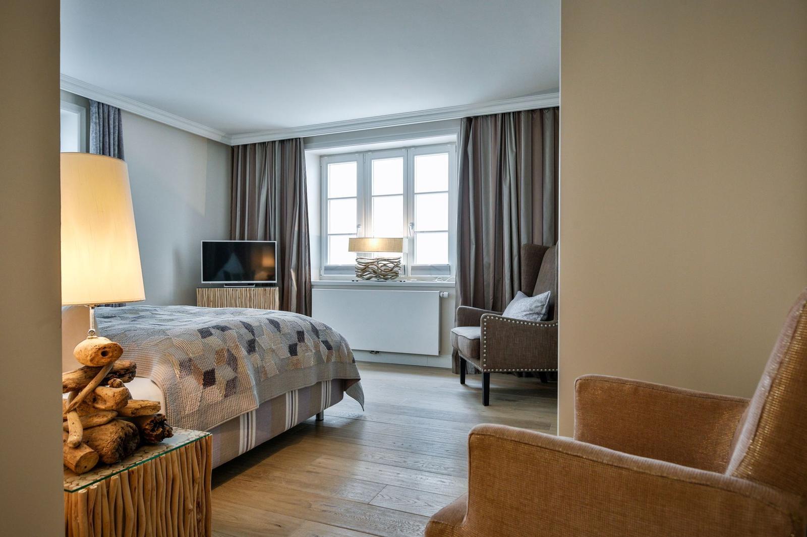 Schlafzimmer mit Bett, TV und Sessel. Holzboden und Fenster mit Vorhängen.