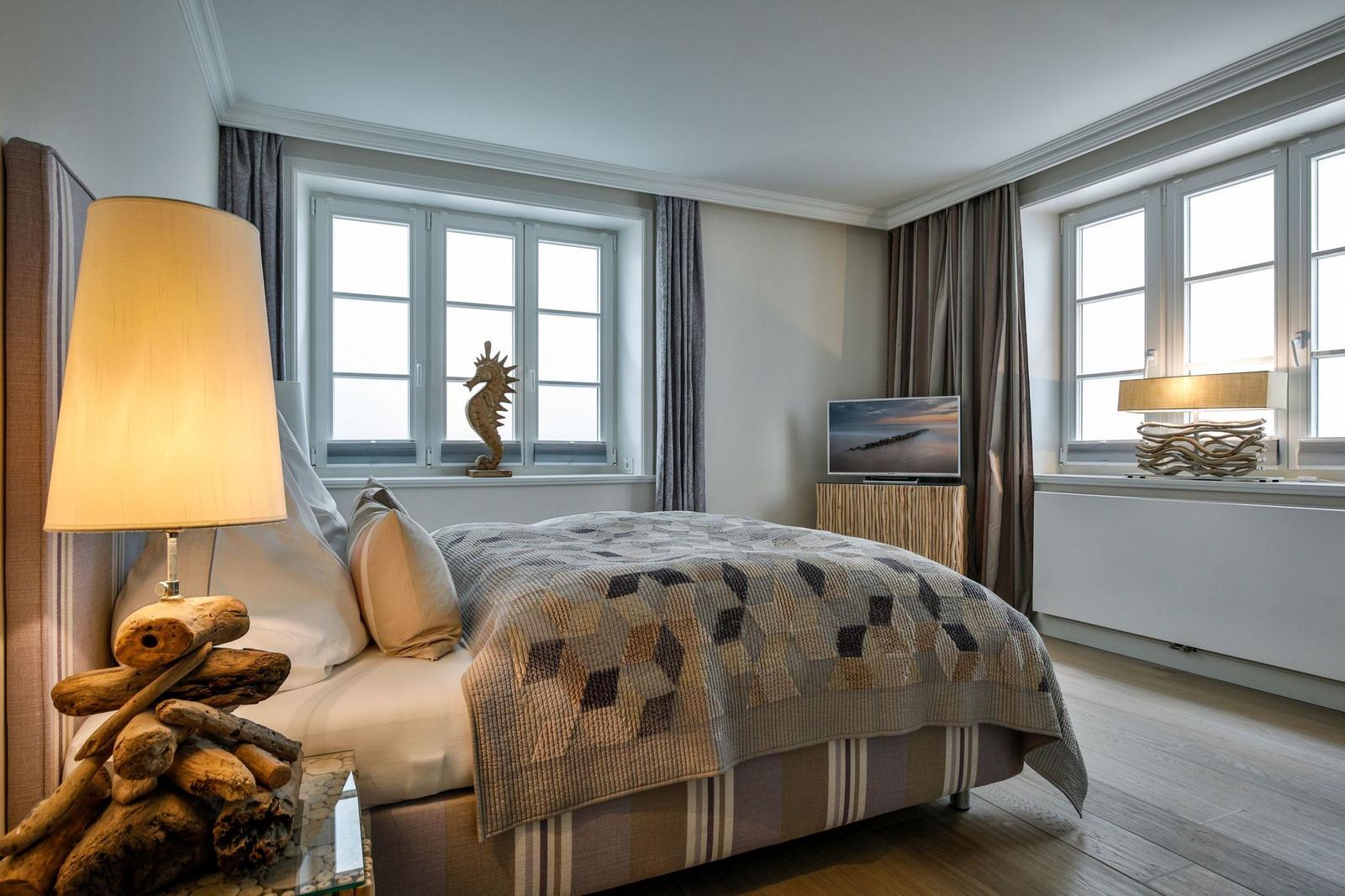 Schlafzimmer mit Bett, Fenstern, Lampe und Fernseher.