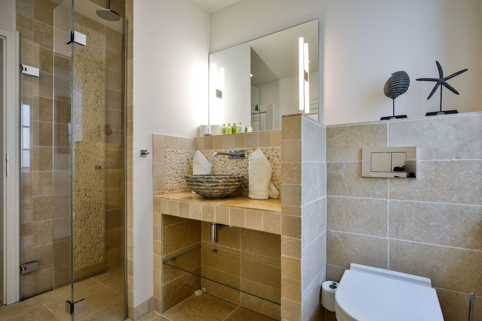 Badezimmer mit Dusche, Waschbecken und Toilette. Steinfliesen und Spiegelschrank.