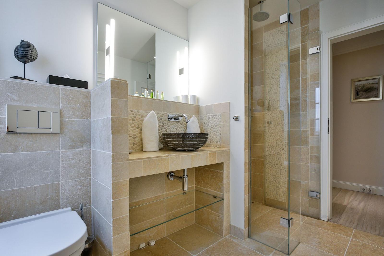 Badezimmer mit Dusche, Waschbecken und Toilette. Steinwand und Spiegel sichtbar.