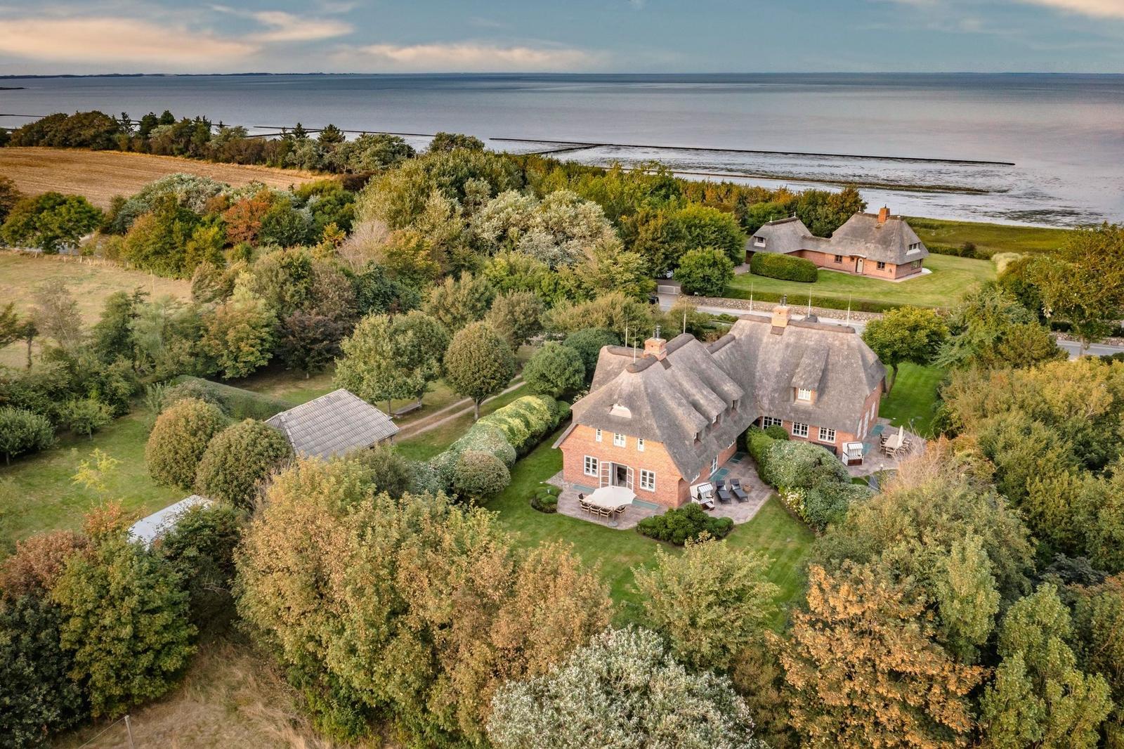 Haus mit Dach aus Stroh, umgeben von Bäumen und Grünflächen, nahe Wasserfläche.