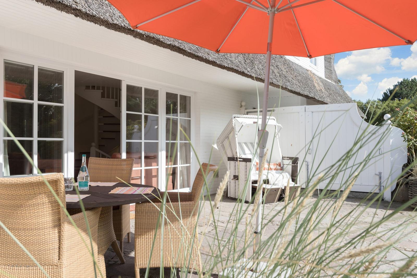 Terrasse mit Tisch, Stühlen und orangefarbenem Sonnenschirm. Hintergrund: weißes Haus mit Schiebetür.
