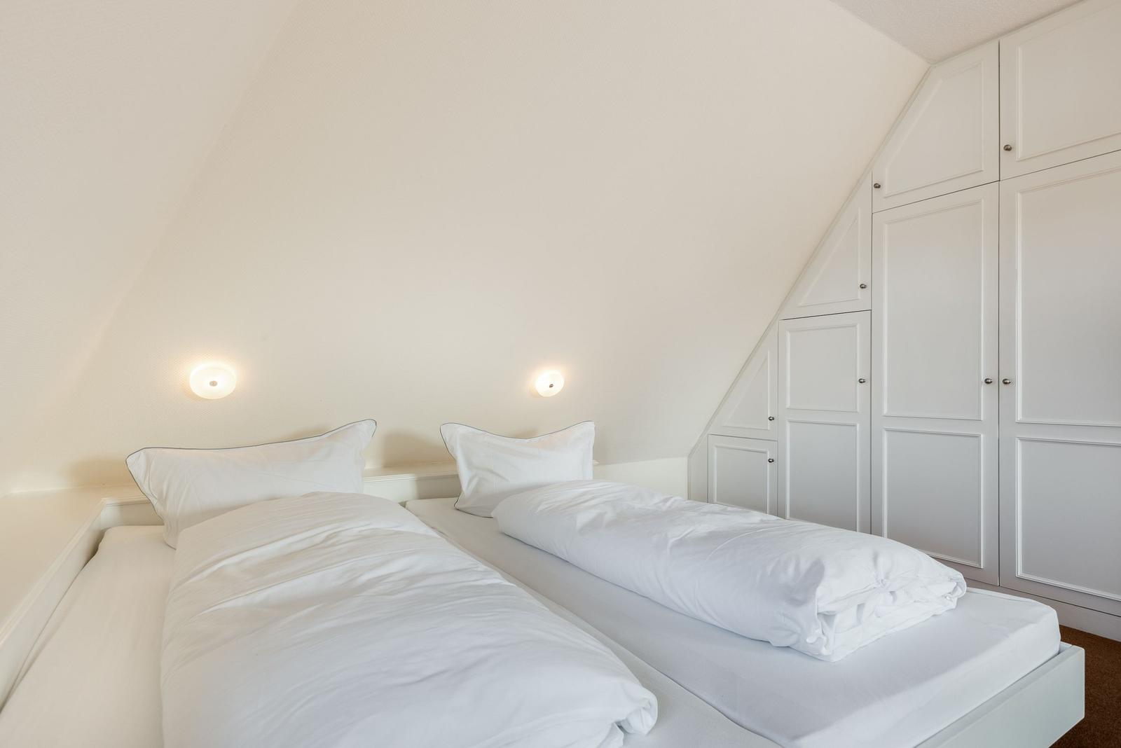 Zwei Betten mit weißen Laken unter einem Dachbodenfenster. Weiße Schränke an der Wand.