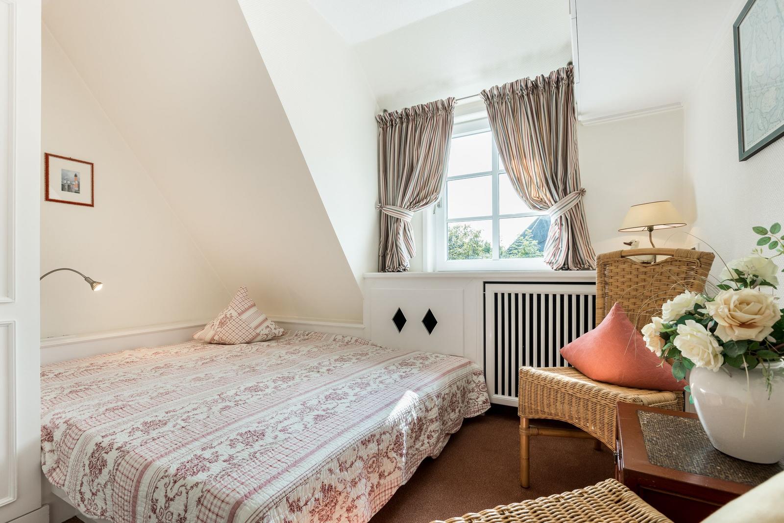 Schlafzimmer mit Dachschrägen, Bett, Fenster und Wickerstuhl.