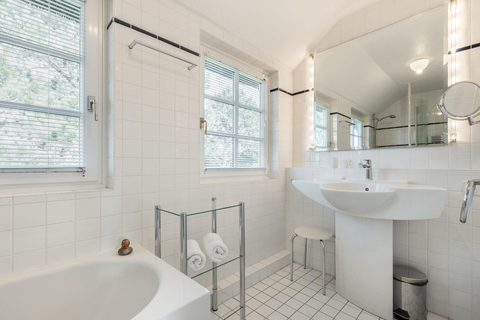 Badezimmer mit Badewanne, Waschbecken und zwei Fenstern mit Jalousien.