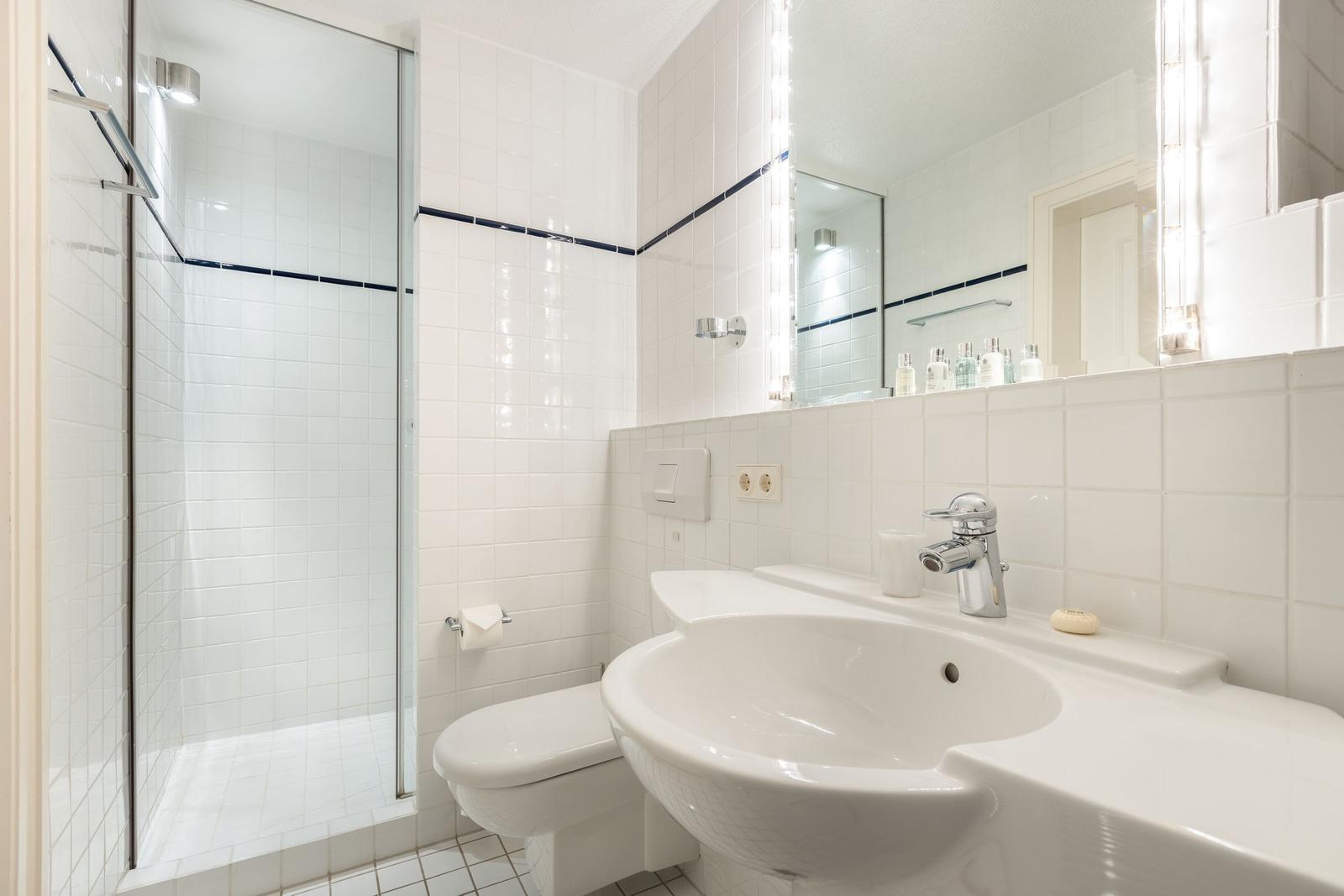 Badezimmer mit Dusche, Waschbecken und Toilette. Weiße Fliesen und Spiegel.
