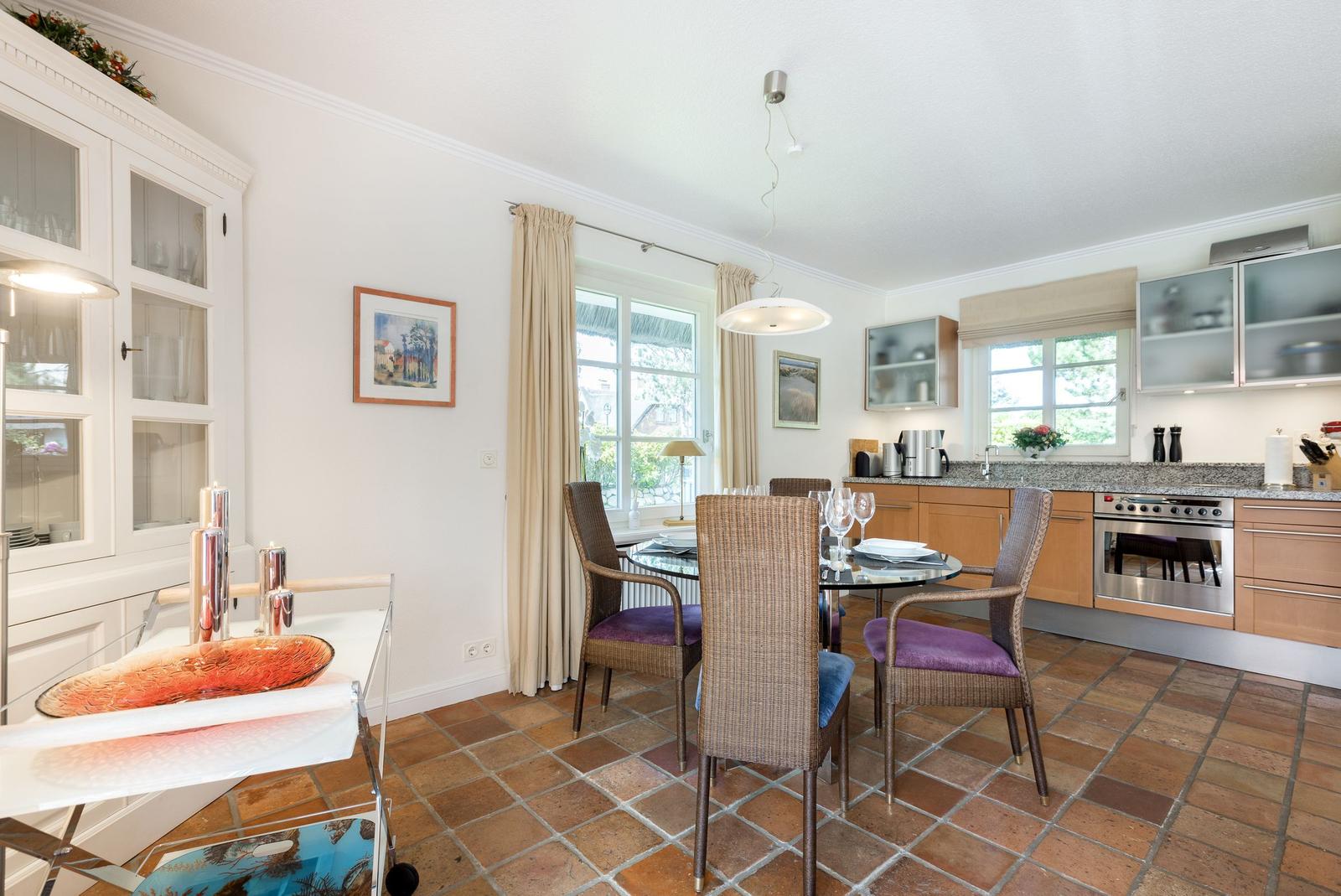 Essbereich mit Tisch, Stühlen und Küchenzeile. Terrakottaboden und Fenster mit Vorhängen.