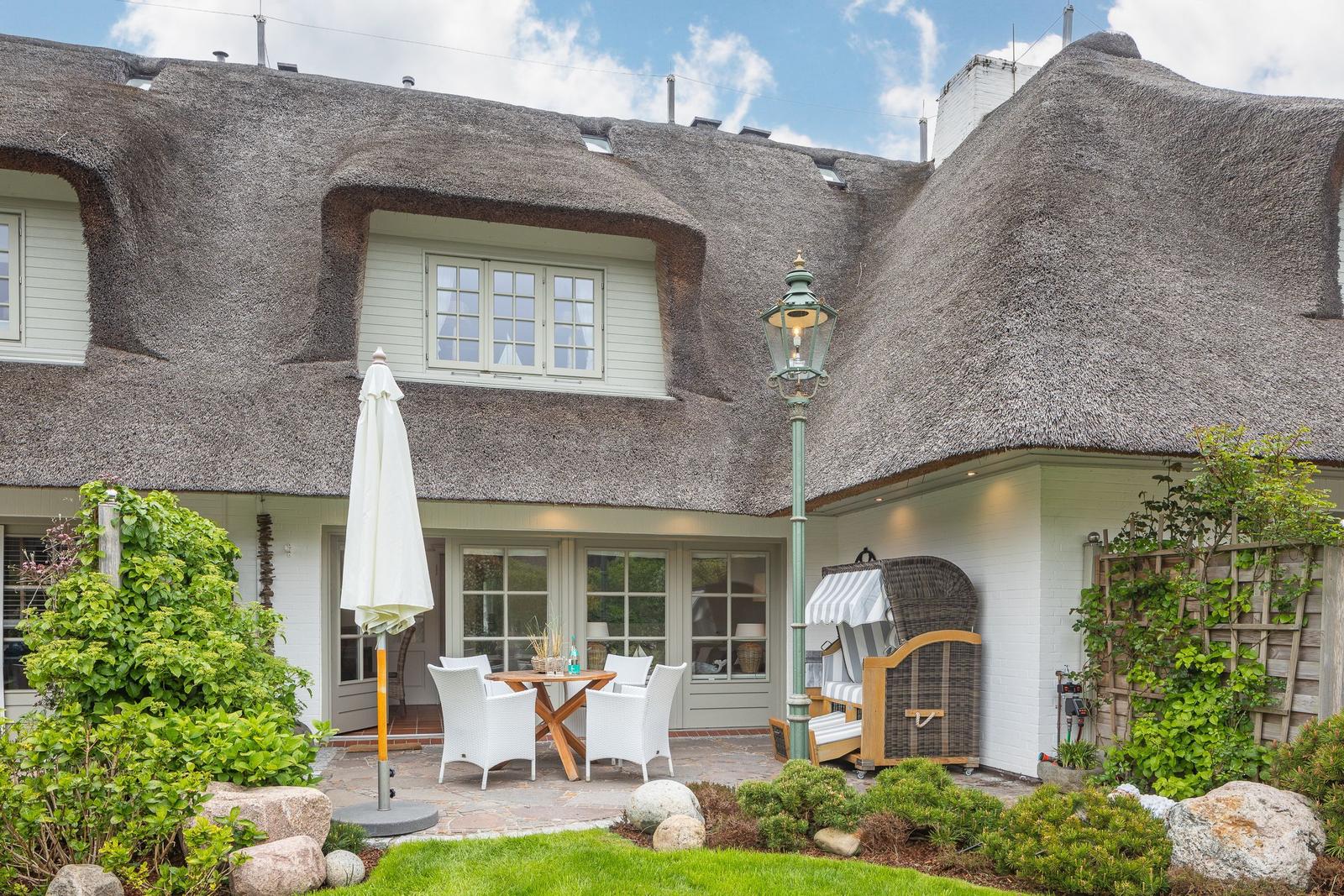 House with thatched roof, terrace with table and chairs, garden with shrubs and stones.