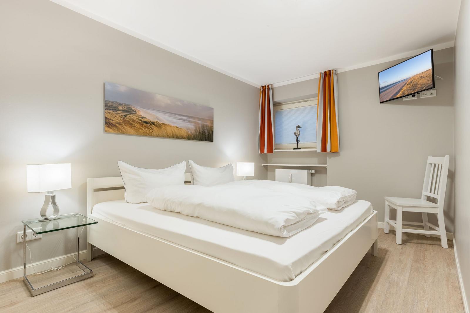Schlafzimmer mit weißem Bett, Fernseher und Fenster mit Vorhängen.