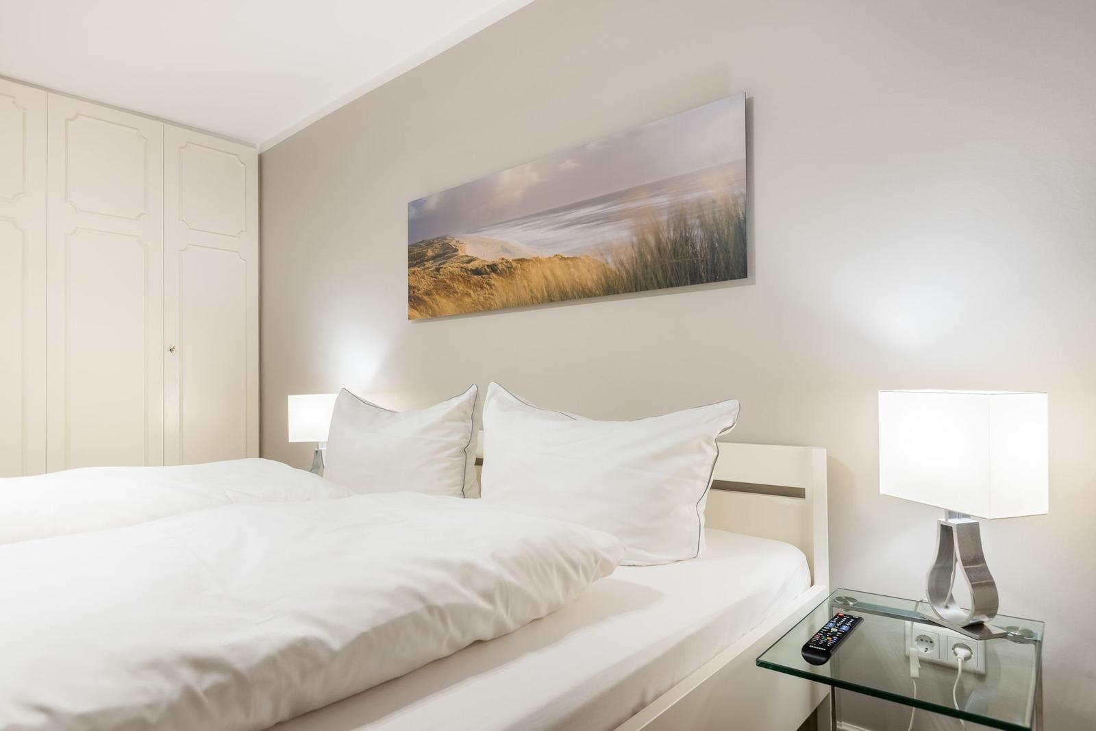 Schlafzimmer mit weißem Bett, Nachttisch und Wandbild.
