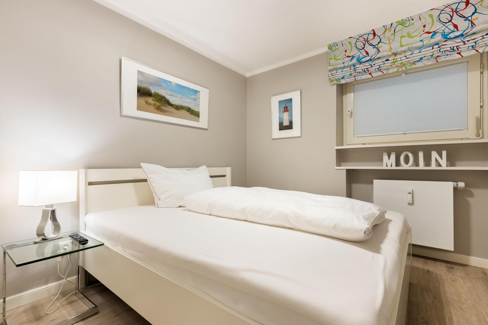 Ein Schlafzimmer mit weißem Bett, Glasnachttisch und buntem Fensterrollo.