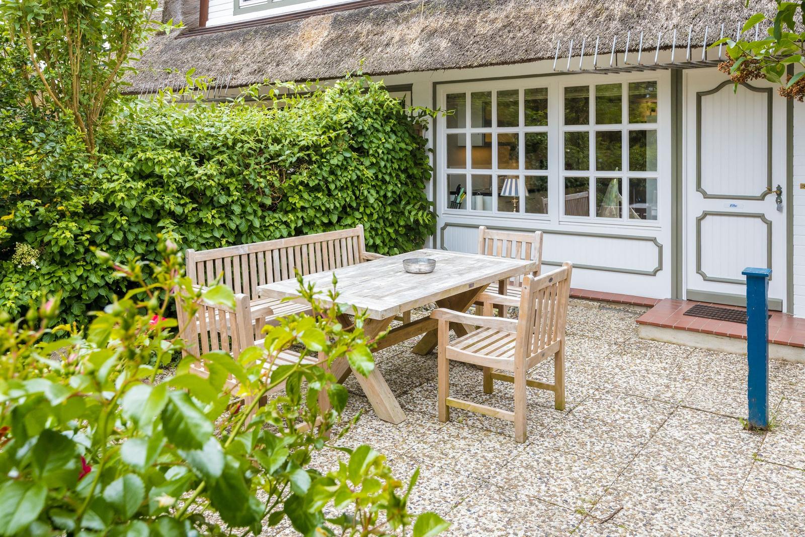 Hinterhof mit Holztisch und Stühlen vor einem Haus mit Strohdach und grünen Hecken.