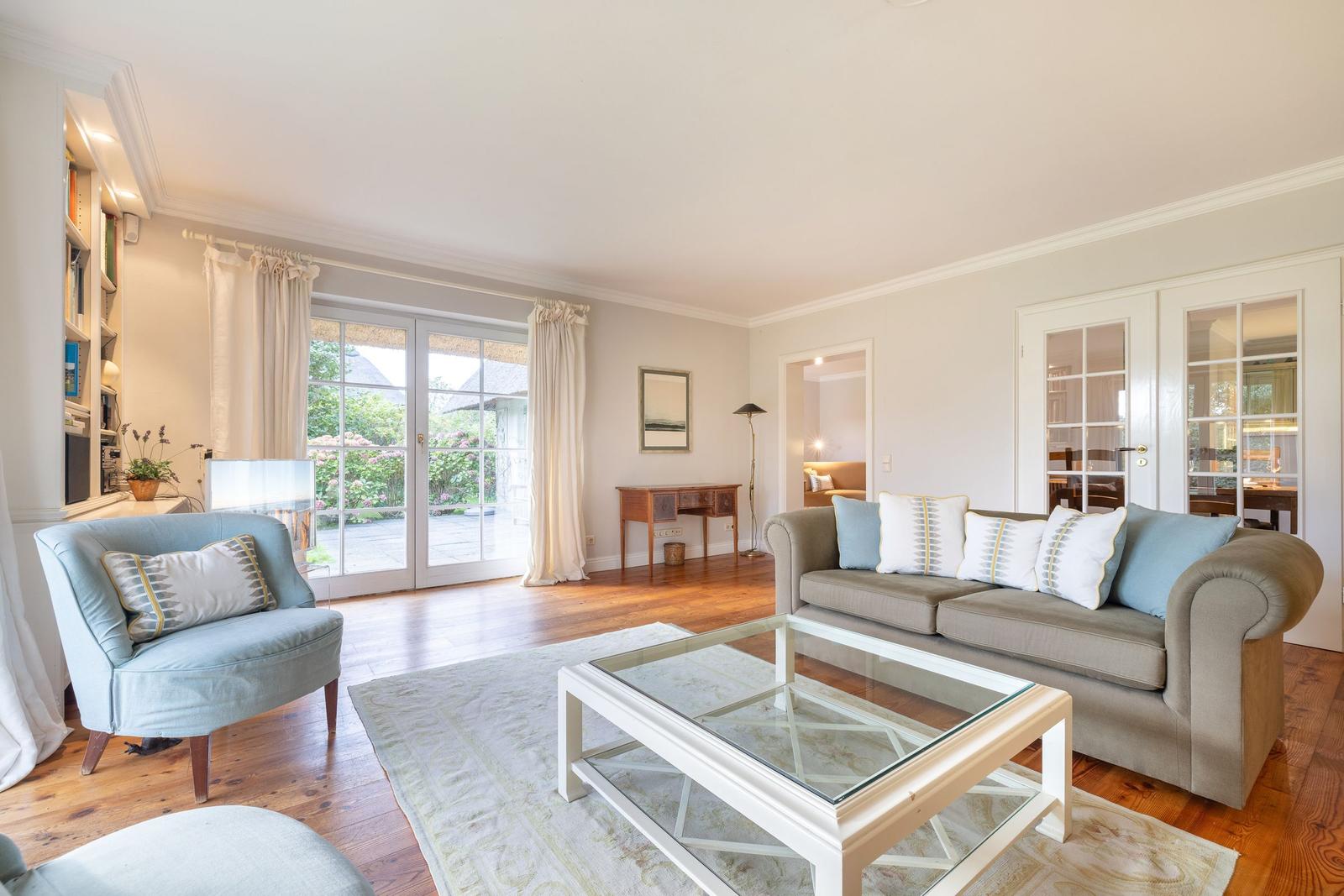 Wohnzimmer mit Sofa, Sessel und Glas-Tisch. Holzboden und Fenster zur Terrasse.