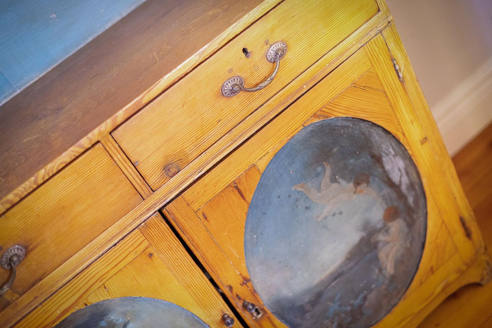 Holzschrank mit zwei Schubladen und runden Fenstern mit Malereien.