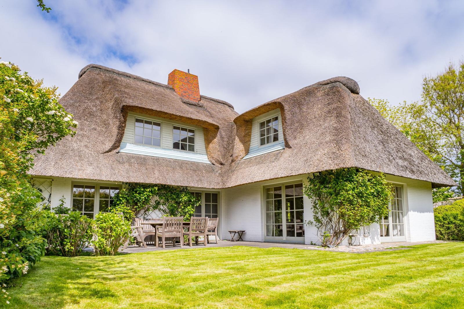 Haus mit Strohdach, Terrasse und Garten. Holztische und Stühle sind draußen platziert.