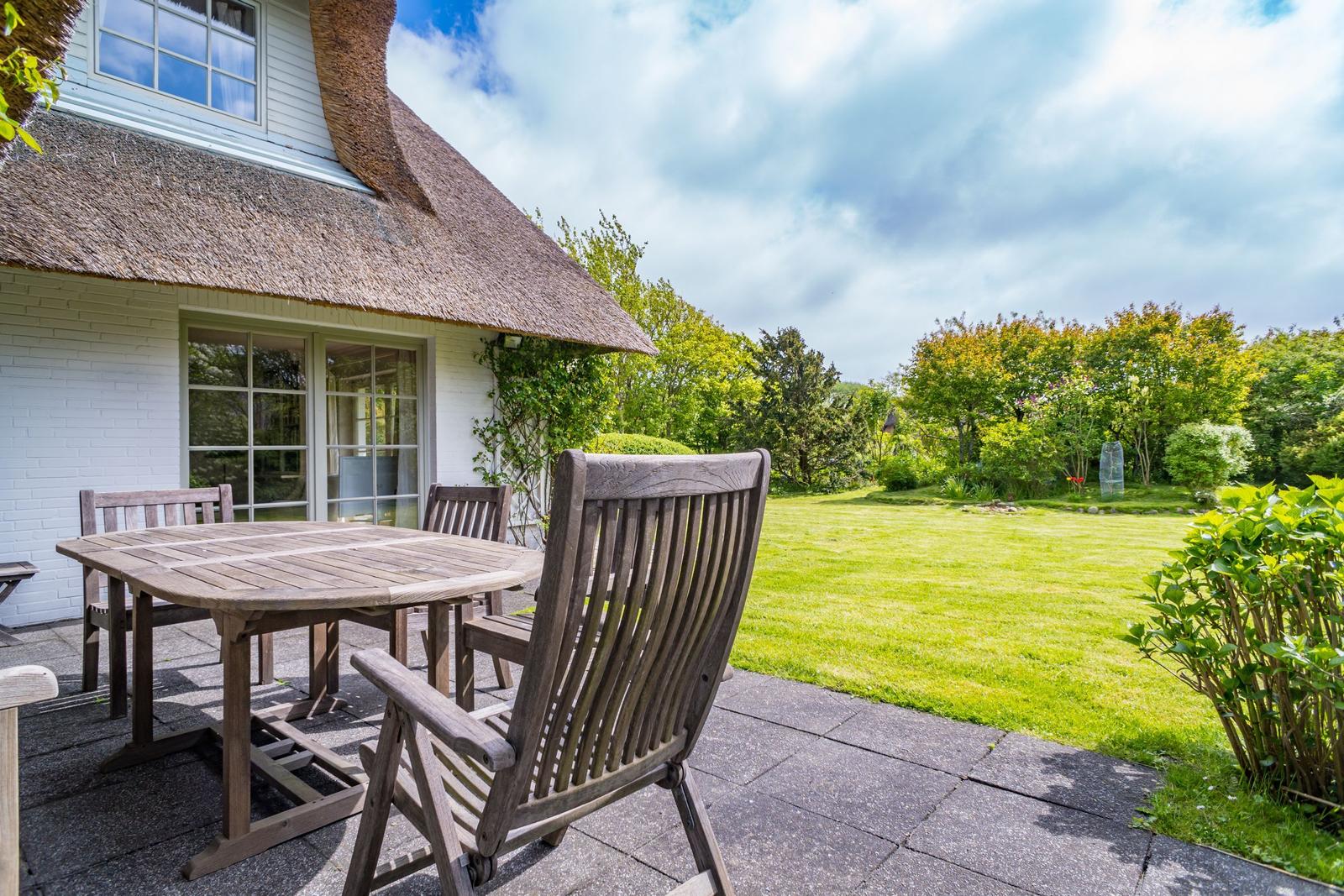 Haus mit Strohdach, Terrasse mit Tisch und Stühlen, grüner Garten im Hintergrund.