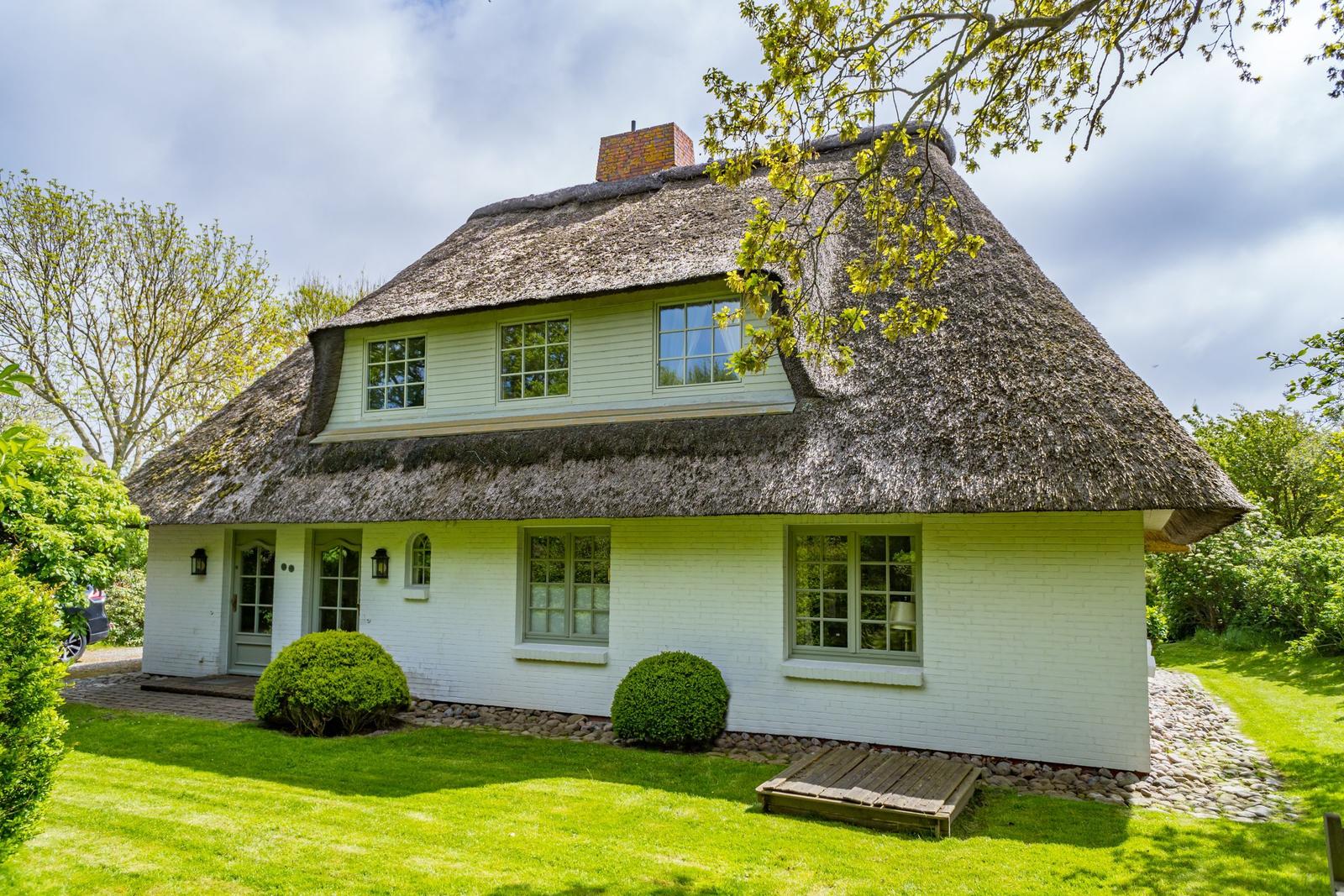 Haus mit strohgedecktem Dach und weißer Fassade, umgeben von Gras und Bäumen.