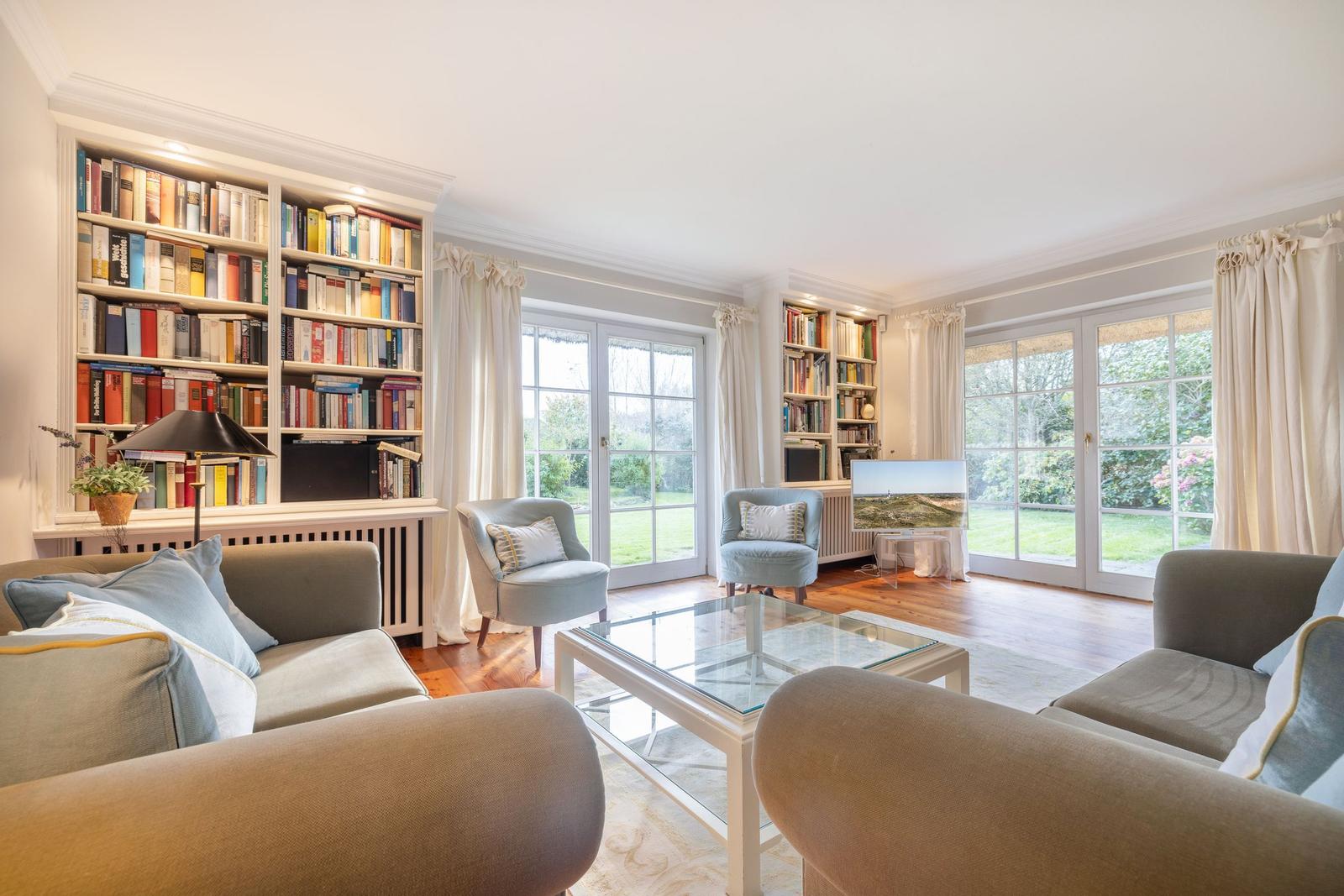 Wohnzimmer mit Bücherregalen, Sesseln und Glas-Tisch. Durchgänge zu Garten.