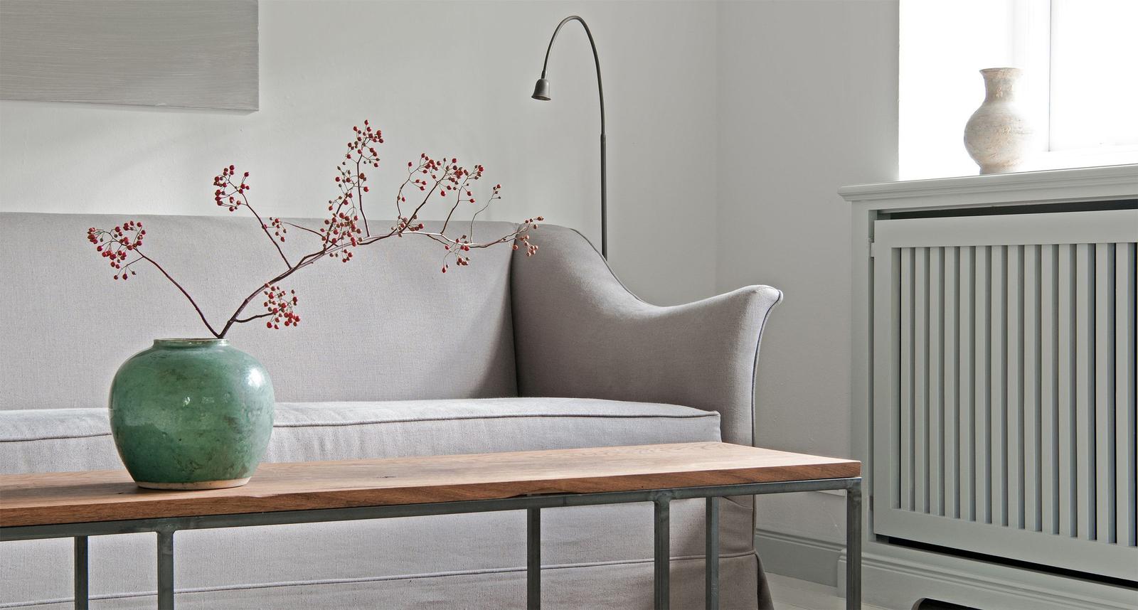 A gray sofa is near a window with a wooden table and a green vase.