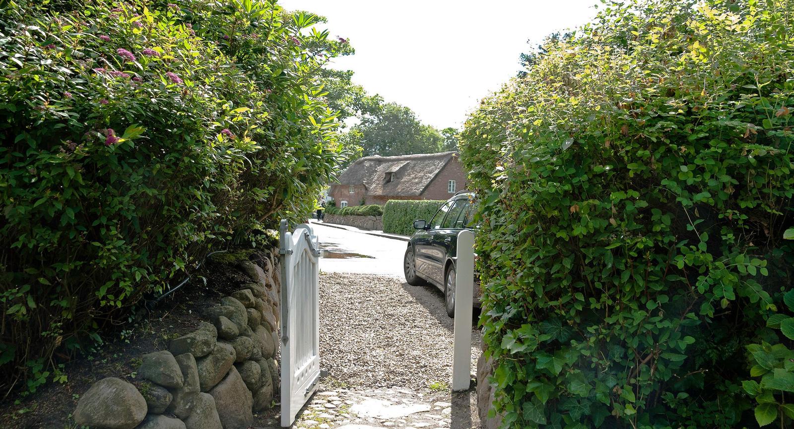 A white gate leads through green hedges to a courtyard with a car and a house in the background.