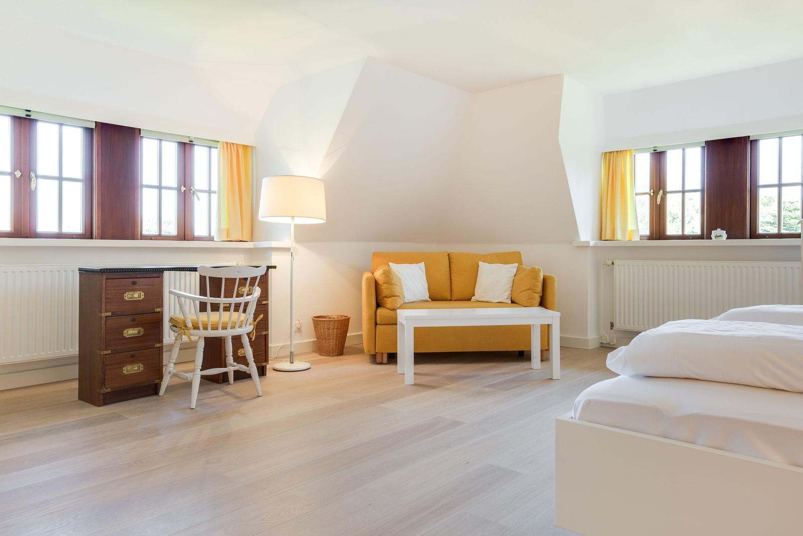 Zimmer mit gelbem Sofa, Schreibtisch und Bett. Holzboden und große Fenster.