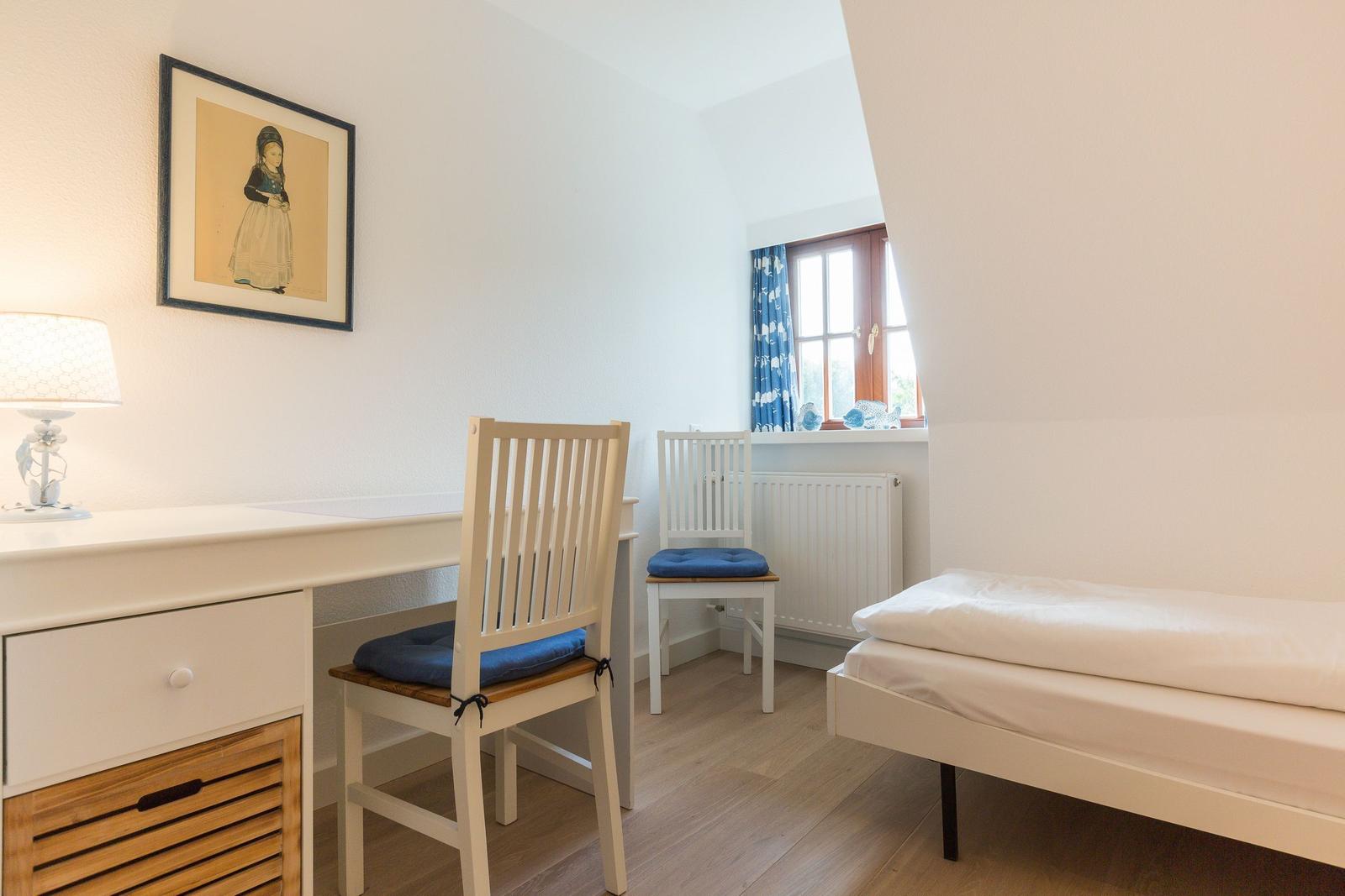 Schlafzimmer mit Schreibtisch, Stühlen und Bett. Fenster mit blauen Vorhängen.