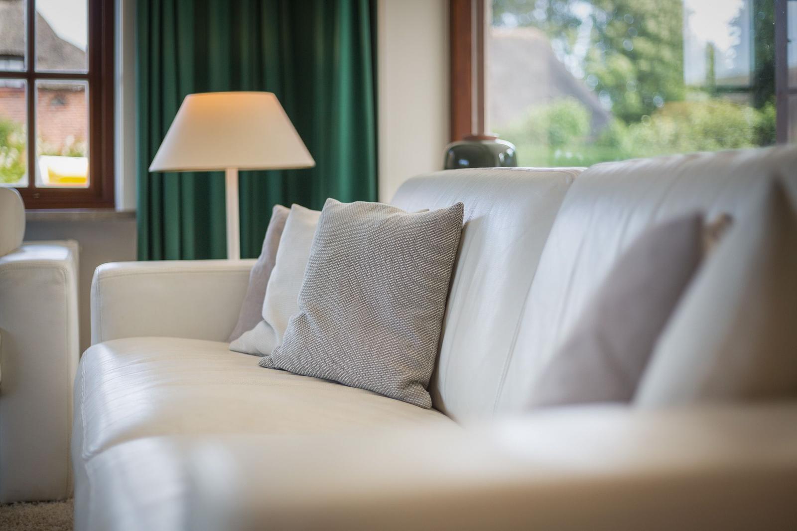 Weißer Ledersofa mit Kissen vor Fenster mit grünen Vorhängen.