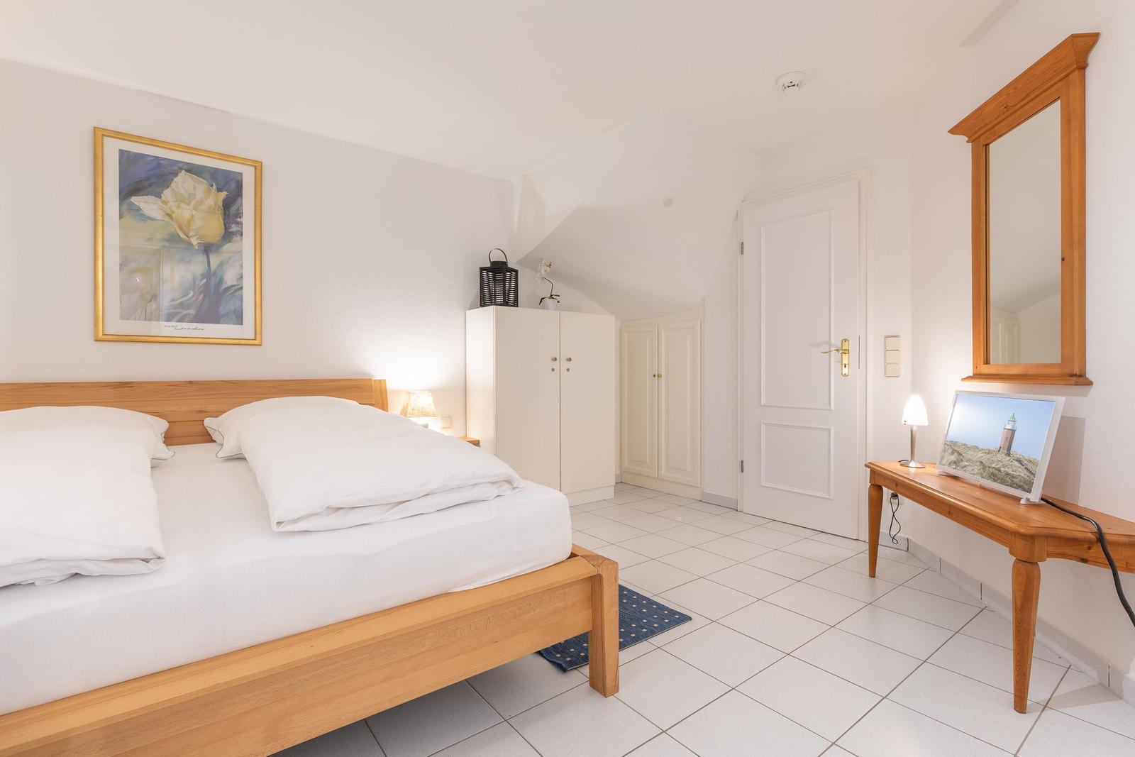 Schlafzimmer mit Holzbett, Schrank und Spiegel. Weiße Wände und Fliesenboden.
