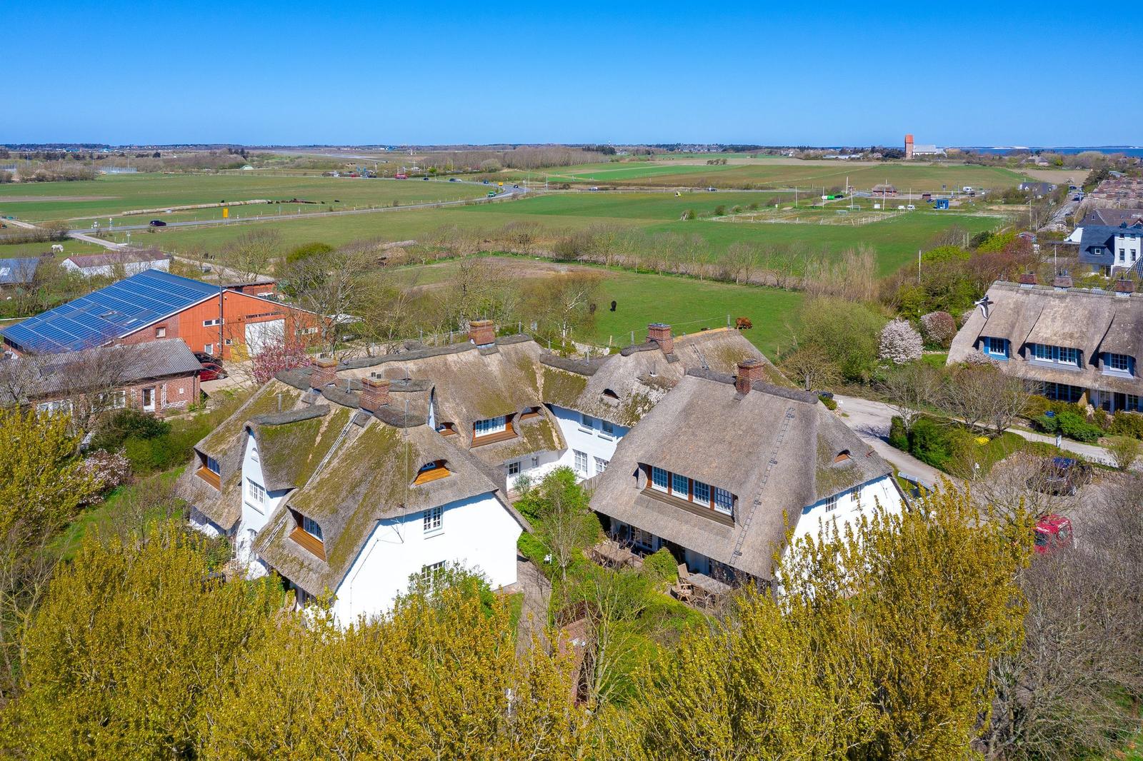 Dorf mit strohgedeckten Häusern und grünen Feldern unter blauem Himmel.
