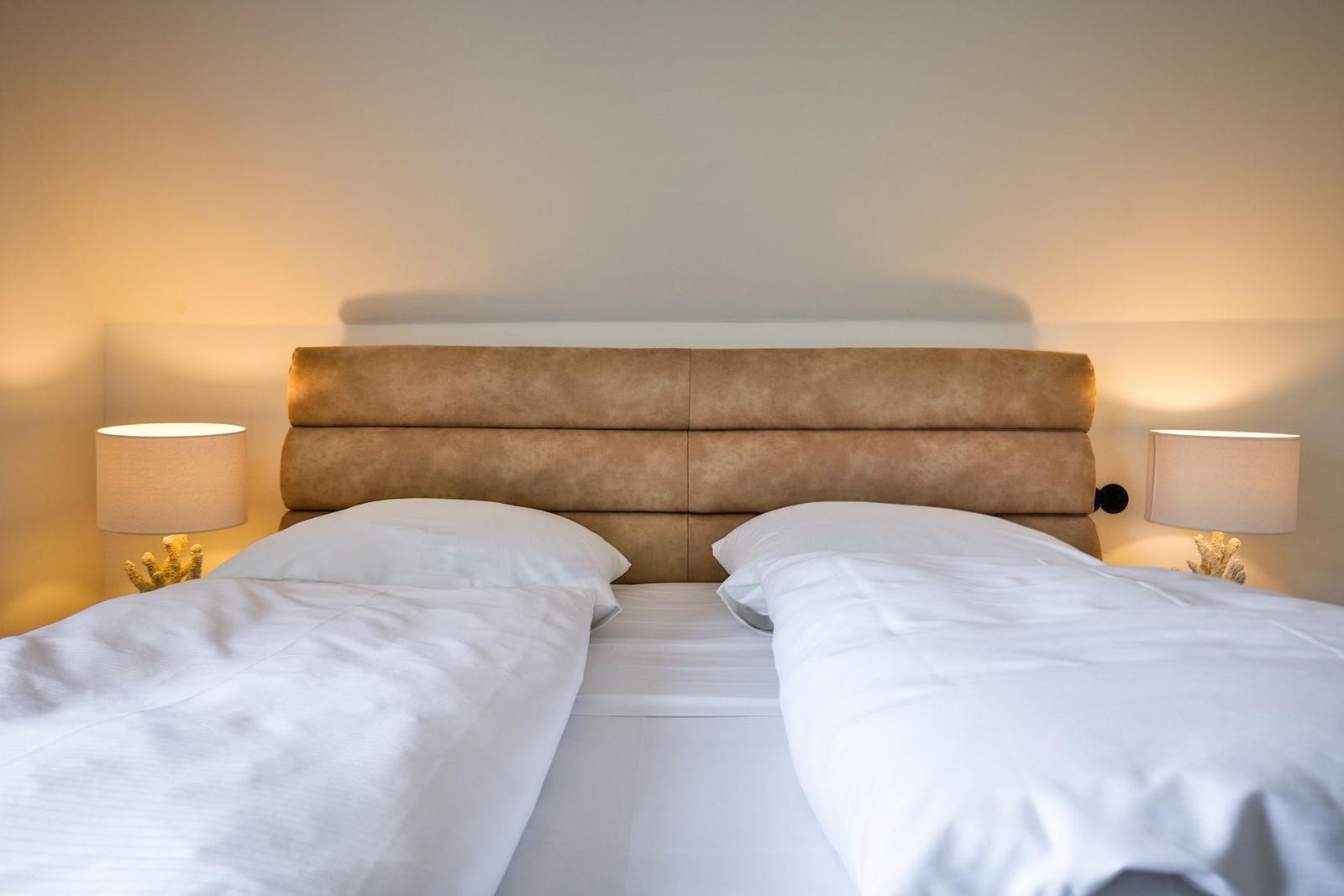 Two beds with white bedding and brown headboard. Both sides have nightstands with lamps.