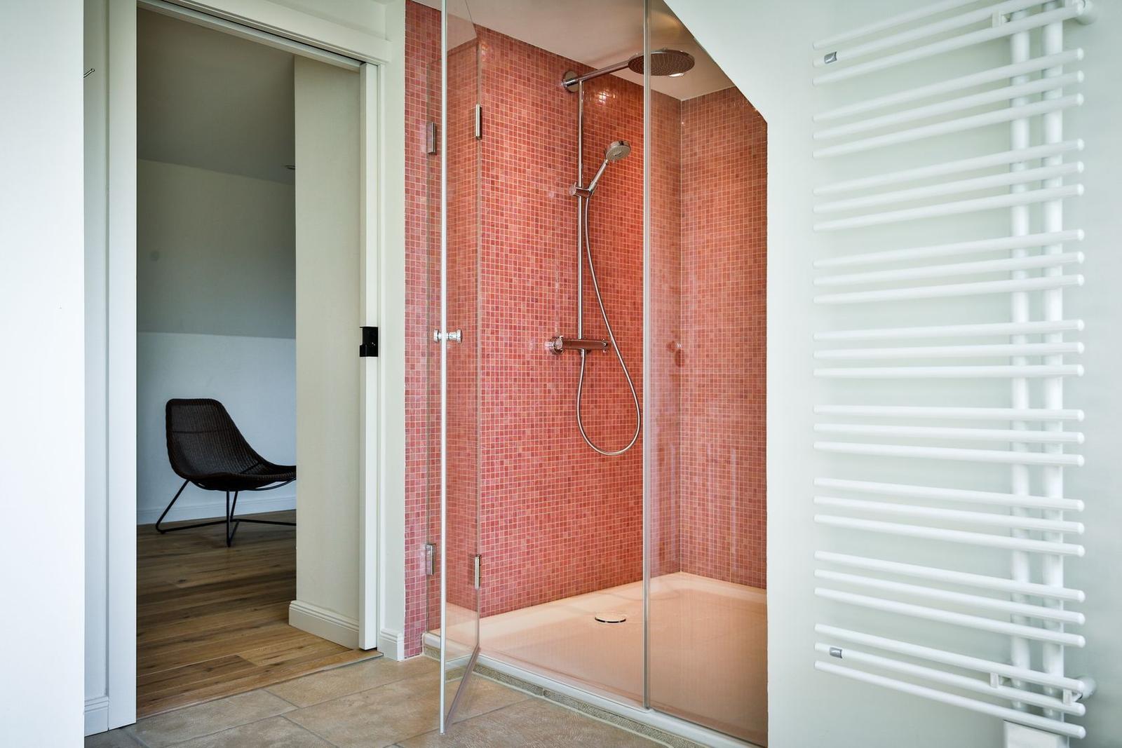 Shower with red tiles and glass enclosure. Radiator on the wall. Door leads to a room with a chair.