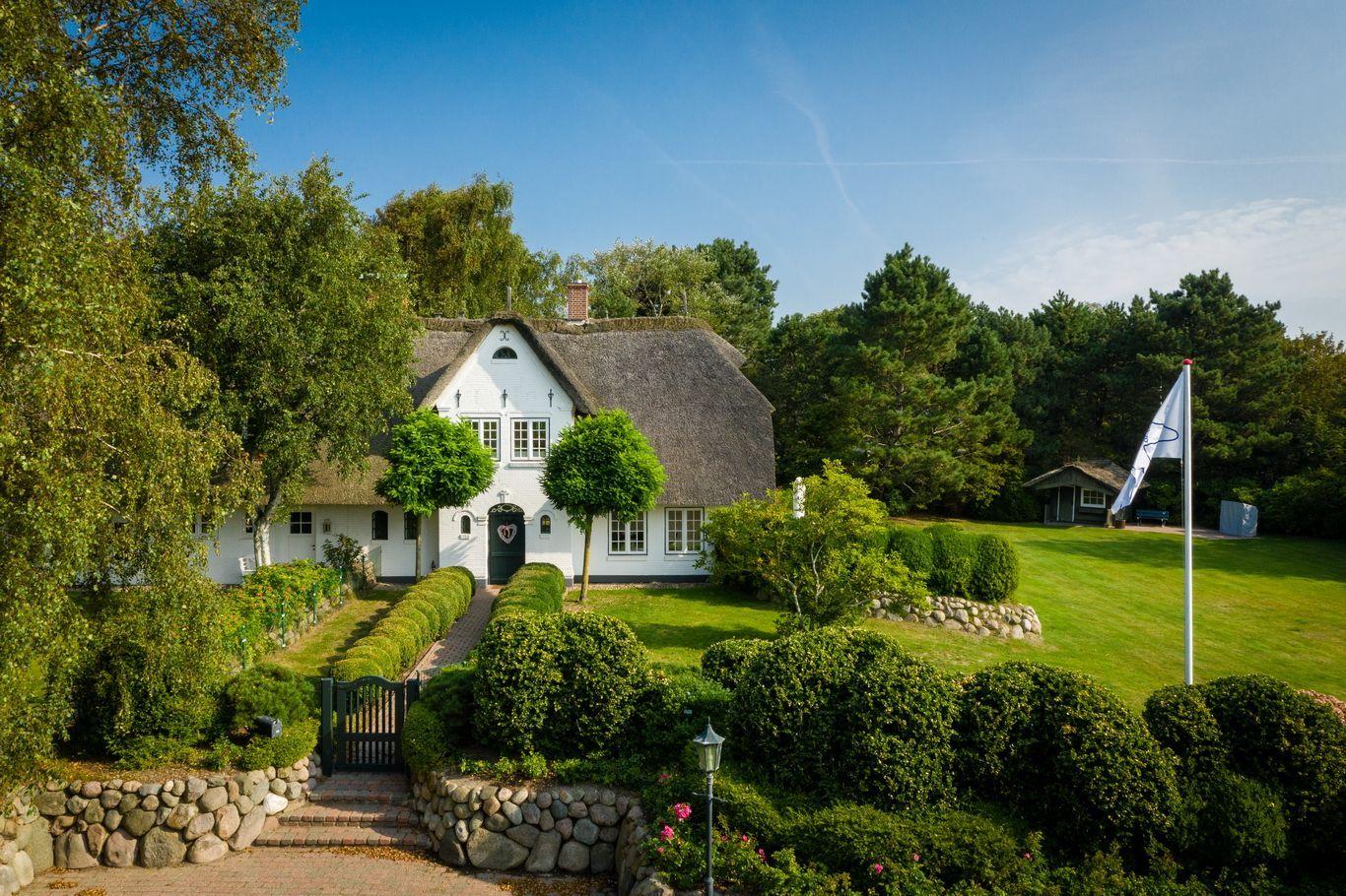 White house with thatched roof, manicured garden, and trees in the background.