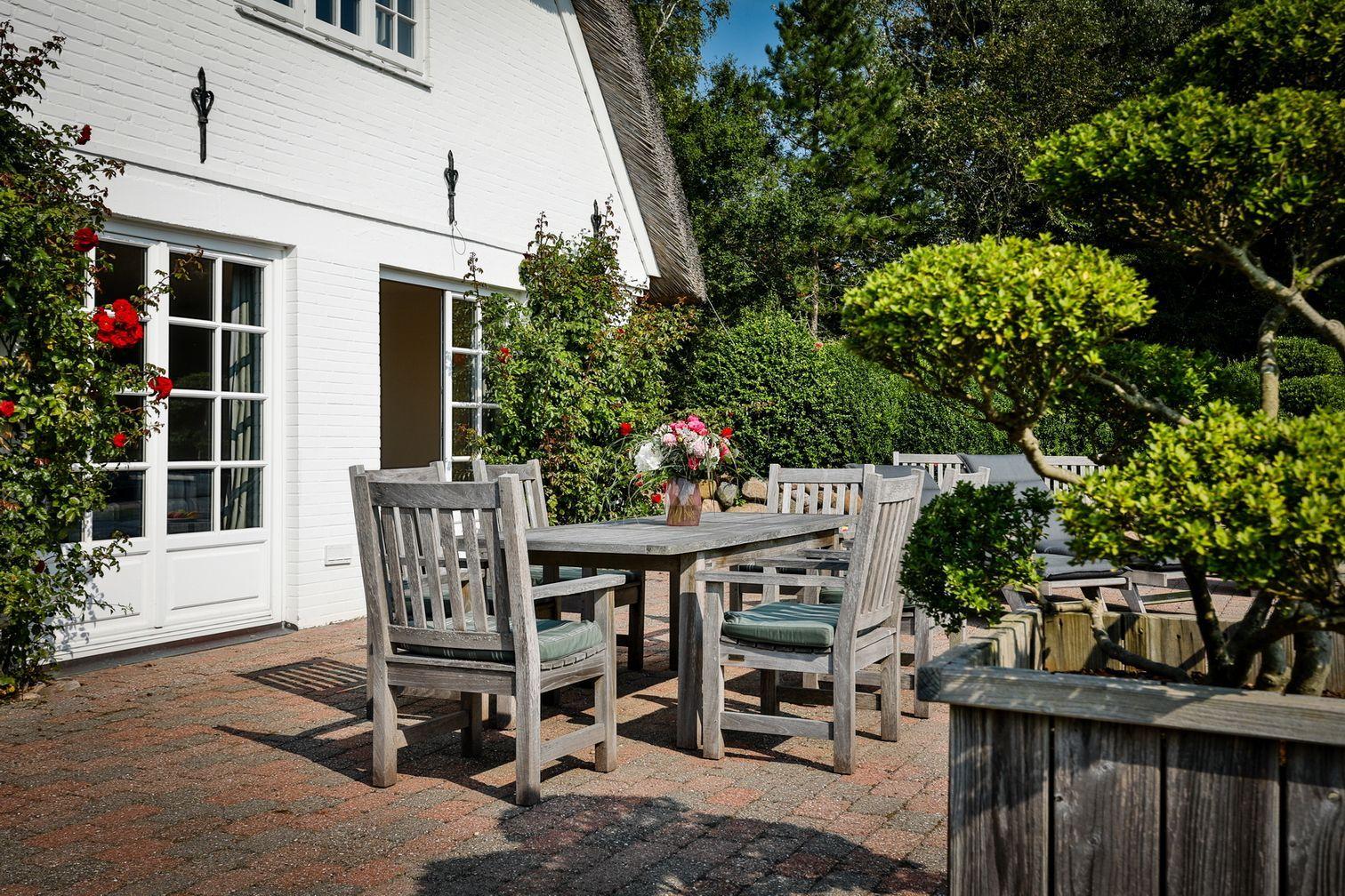 Bright terrace with wooden table, chairs, and flower arrangement. White house wall and green plants in background.