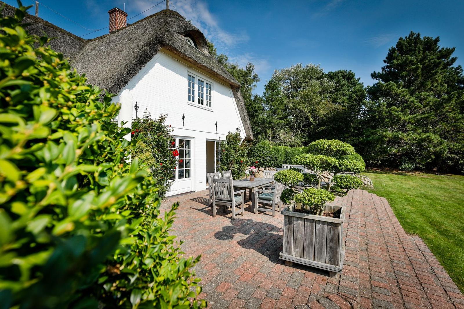 White house with thatched roof, terrace with table and chairs, landscaped garden with trees.