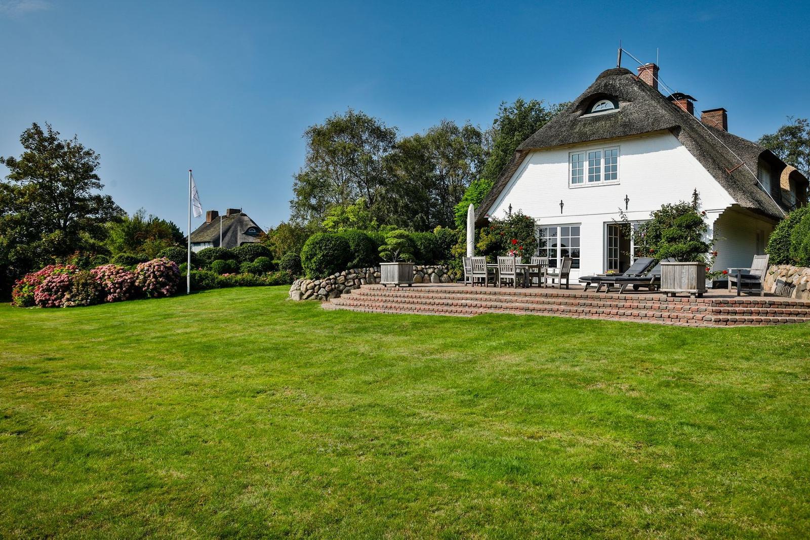 House with thatched roof, terrace, and garden. Wooden furniture and flowers are visible.